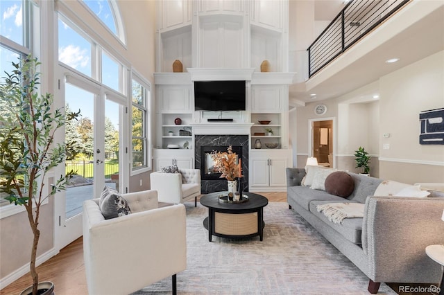 living room with built in features, light wood-type flooring, a premium fireplace, a towering ceiling, and french doors