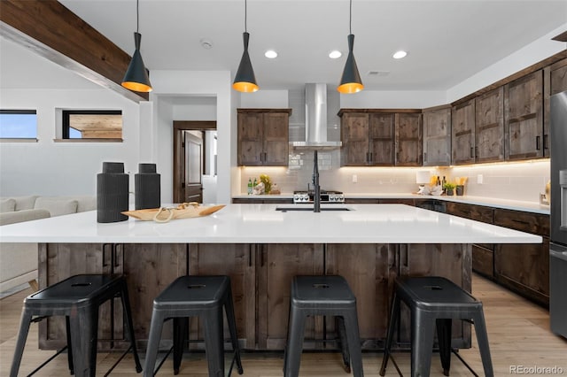 kitchen with wall chimney range hood, light wood finished floors, decorative backsplash, dark brown cabinets, and light countertops