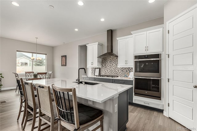 kitchen featuring backsplash, wall chimney range hood, a sink, light wood-style floors, and appliances with stainless steel finishes