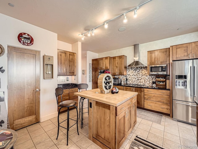 kitchen with wall chimney exhaust hood, butcher block countertops, appliances with stainless steel finishes, a kitchen breakfast bar, and backsplash