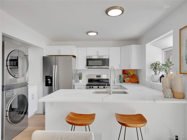 kitchen featuring stainless steel appliances, stacked washer and clothes dryer, a peninsula, a sink, and white cabinetry
