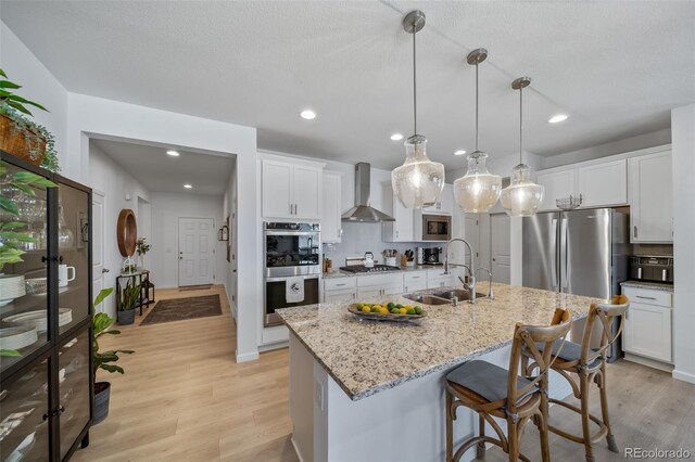 kitchen with wall chimney range hood, a sink, light wood-style floors, stainless steel appliances, and white cabinetry