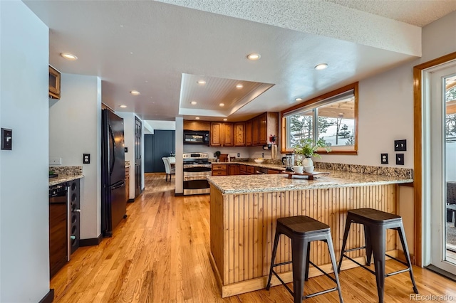 kitchen featuring a tray ceiling, brown cabinetry, black appliances, a peninsula, and a kitchen bar