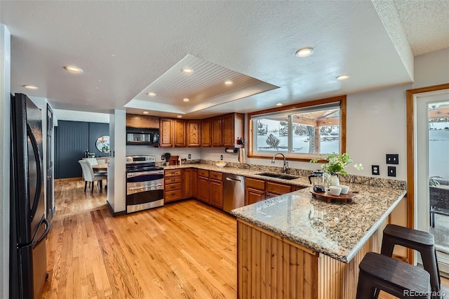 kitchen featuring brown cabinetry, a raised ceiling, a peninsula, black appliances, and a sink