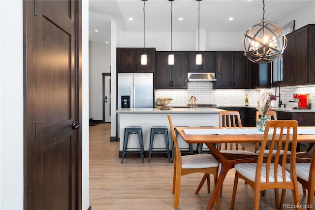 kitchen featuring decorative light fixtures, backsplash, stainless steel fridge, dark brown cabinetry, and light wood-type flooring