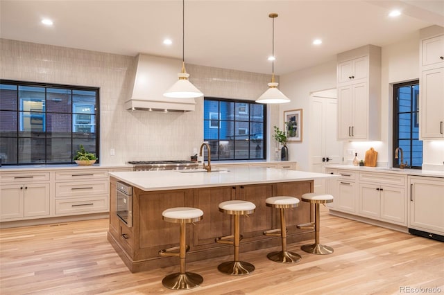 kitchen with a center island with sink, light countertops, a sink, light wood-type flooring, and premium range hood