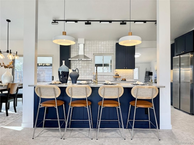 kitchen featuring stainless steel refrigerator, a kitchen bar, hanging light fixtures, and wall chimney range hood