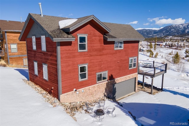 snow covered property featuring a fire pit, roof with shingles, an attached garage, and a mountain view