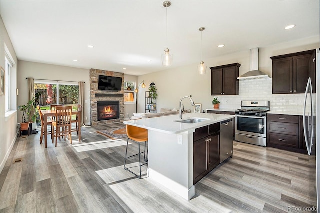 kitchen featuring stainless steel appliances, sink, wall chimney range hood, decorative light fixtures, and a center island with sink