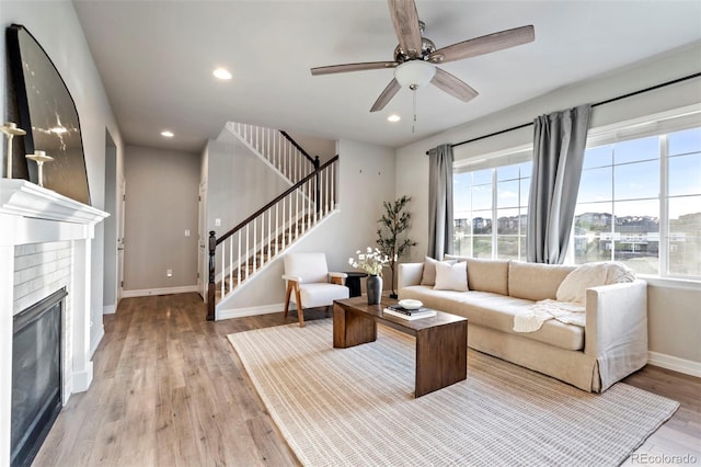 living room featuring a brick fireplace, stairs, a ceiling fan, baseboards, and wood finished floors