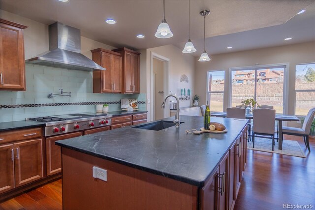 kitchen with sink, hanging light fixtures, wall chimney exhaust hood, an island with sink, and tasteful backsplash