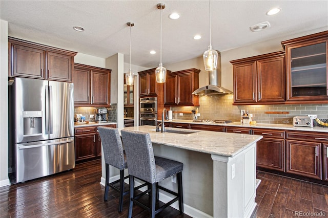 kitchen with dark wood finished floors, appliances with stainless steel finishes, a kitchen island with sink, wall chimney range hood, and a sink