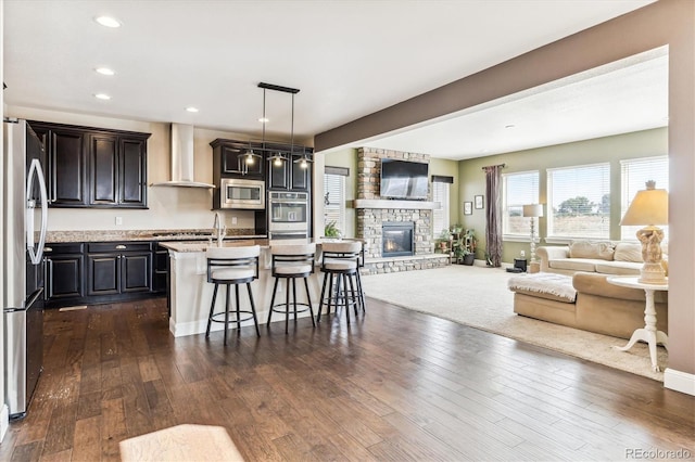 kitchen featuring pendant lighting, stainless steel appliances, an island with sink, a kitchen bar, and wall chimney exhaust hood