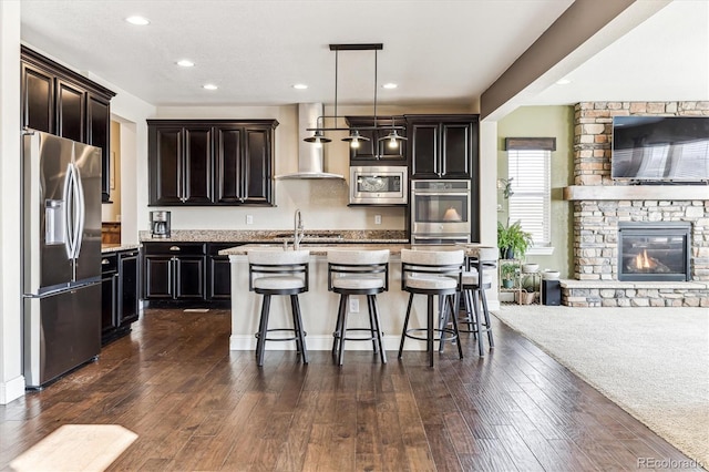 kitchen with a kitchen bar, pendant lighting, stainless steel appliances, a kitchen island with sink, and wall chimney range hood