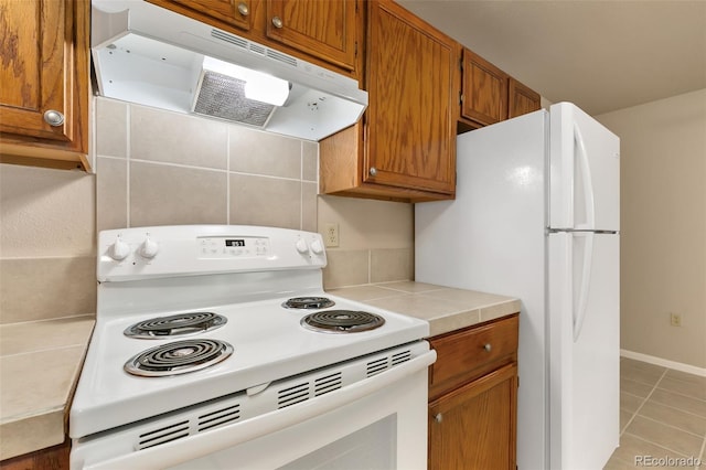kitchen with brown cabinets, light tile patterned floors, under cabinet range hood, and white appliances