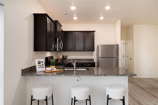 kitchen with a peninsula, a sink, dark brown cabinets, appliances with stainless steel finishes, and dark stone counters