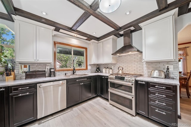 kitchen with appliances with stainless steel finishes, wall chimney range hood, a sink, white cabinets, and beam ceiling