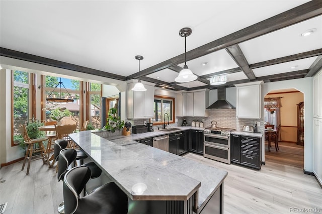 kitchen featuring stainless steel appliances, wall chimney exhaust hood, a peninsula, light wood-style flooring, and beamed ceiling