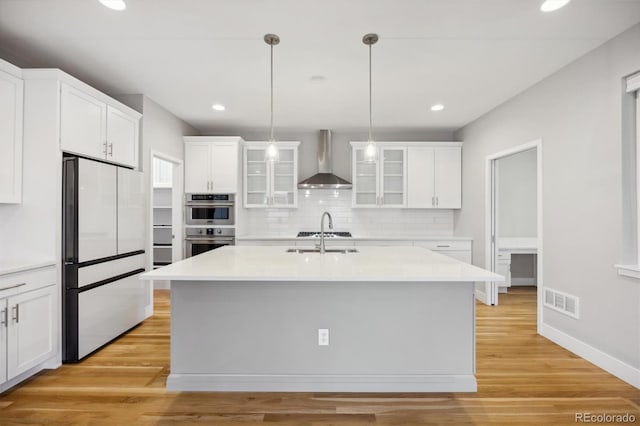 kitchen with white cabinetry, a center island with sink, wall chimney range hood, and appliances with stainless steel finishes