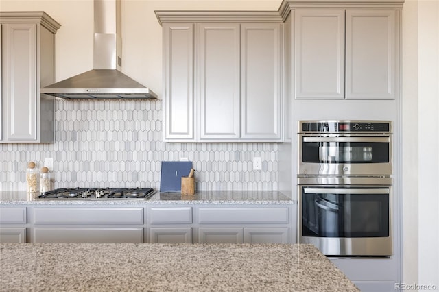 kitchen with wall chimney exhaust hood, light stone counters, decorative backsplash, and appliances with stainless steel finishes