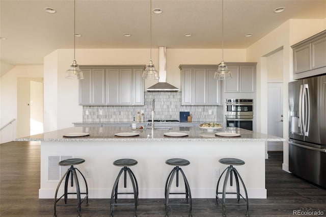 kitchen with pendant lighting, wall chimney exhaust hood, stainless steel appliances, and a kitchen island with sink