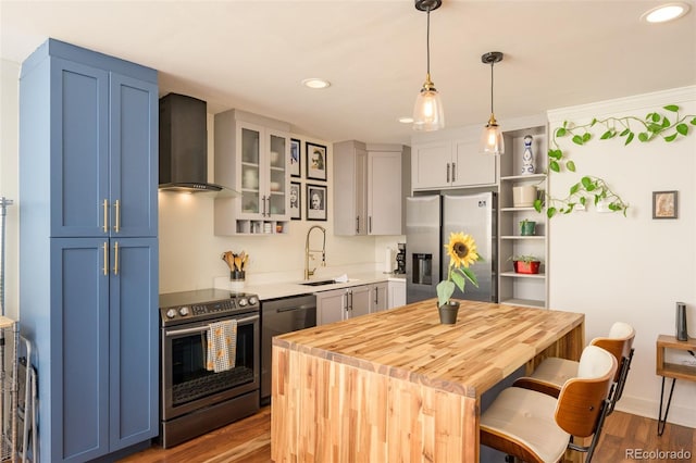 kitchen with wall chimney range hood, wood-type flooring, sink, stainless steel appliances, and blue cabinets