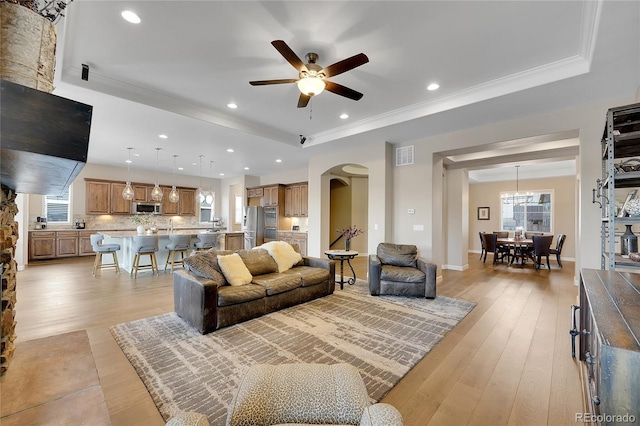 living room featuring a tray ceiling, arched walkways, a ceiling fan, light wood finished floors, and crown molding