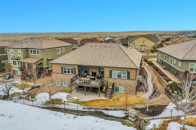 snow covered back of property with a deck, stone siding, a fenced backyard, and a residential view