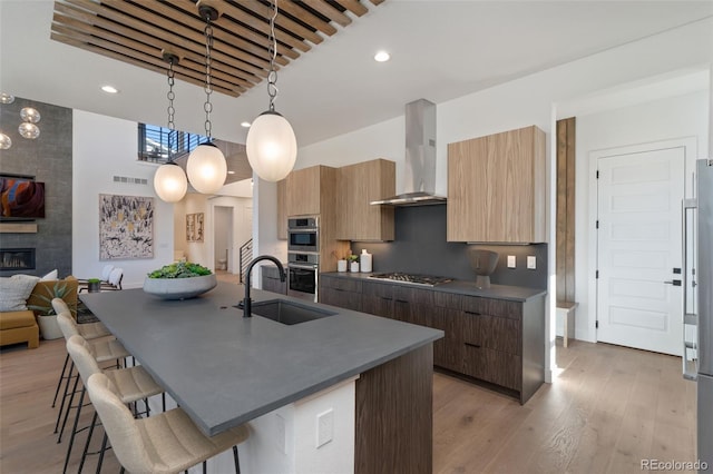 kitchen featuring sink, an island with sink, a tile fireplace, pendant lighting, and wall chimney range hood