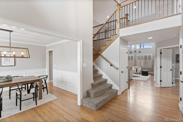 stairs with an inviting chandelier, wood finished floors, a wainscoted wall, crown molding, and a towering ceiling
