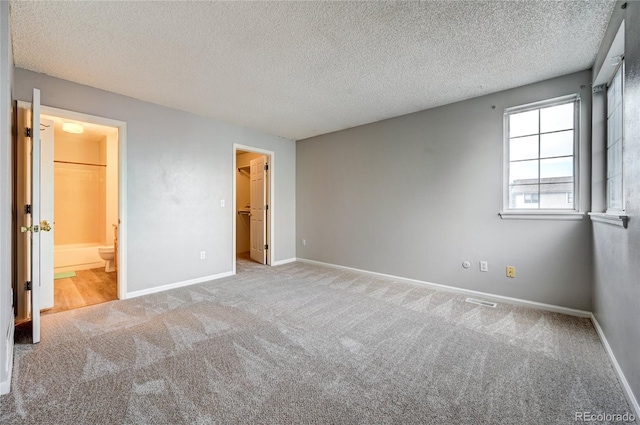 unfurnished bedroom featuring ensuite bathroom, a walk in closet, light carpet, and a textured ceiling