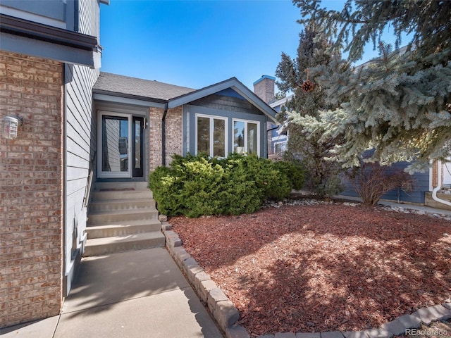 doorway to property featuring a shingled roof, brick siding, and a chimney