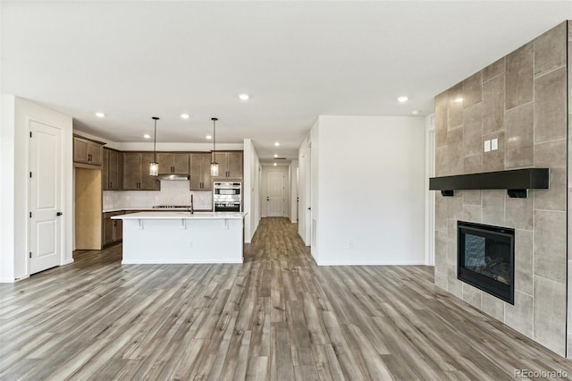 kitchen with stainless steel double oven, light hardwood / wood-style flooring, a center island with sink, pendant lighting, and a fireplace