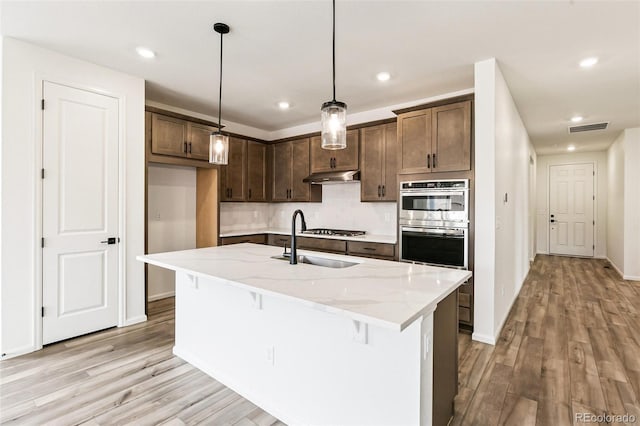 kitchen featuring an island with sink, sink, pendant lighting, light wood-type flooring, and light stone counters