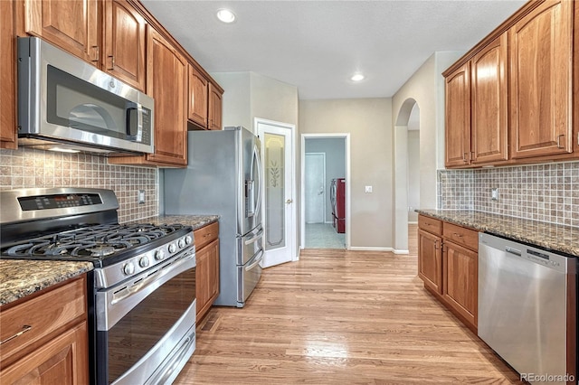 kitchen featuring light wood-style flooring, dark stone countertops, arched walkways, appliances with stainless steel finishes, and brown cabinetry