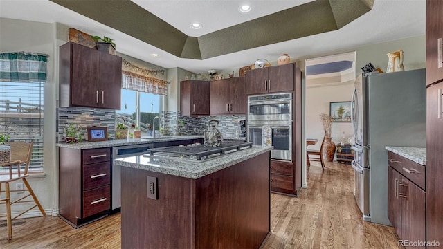 kitchen with a tray ceiling, a sink, light wood finished floors, backsplash, and stainless steel appliances
