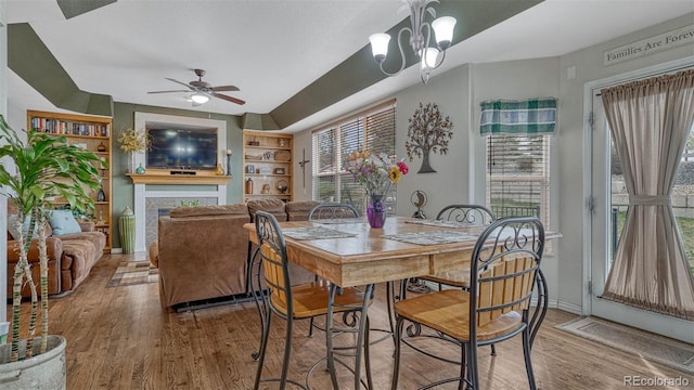 dining area with a tiled fireplace, ceiling fan with notable chandelier, wood finished floors, and a healthy amount of sunlight