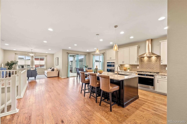 kitchen with sink, white cabinetry, pendant lighting, wall chimney range hood, and a kitchen island with sink