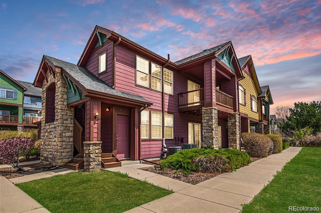 view of front facade featuring a balcony, central AC, stone siding, roof with shingles, and a front yard