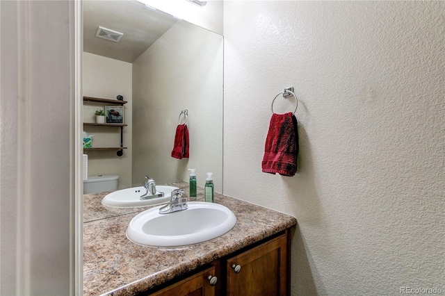 half bathroom with vanity, toilet, a textured wall, and visible vents