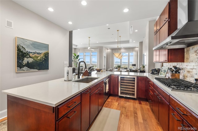 kitchen featuring a sink, wall chimney range hood, light wood-type flooring, stainless steel gas stovetop, and beverage cooler