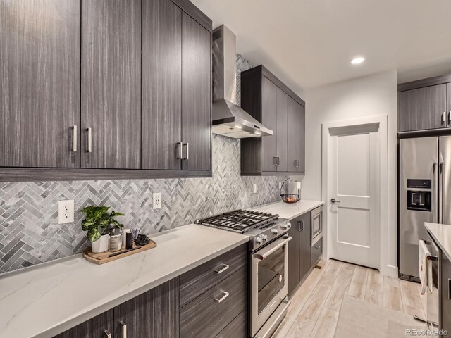 kitchen featuring wall chimney exhaust hood, dark brown cabinets, appliances with stainless steel finishes, and light hardwood / wood-style flooring