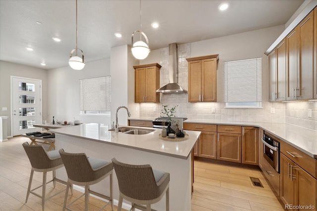 kitchen featuring sink, wall chimney range hood, oven, decorative light fixtures, and a kitchen island with sink