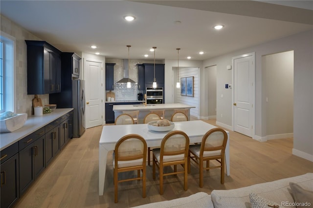 kitchen featuring wall chimney exhaust hood, decorative light fixtures, a kitchen island with sink, and light hardwood / wood-style flooring