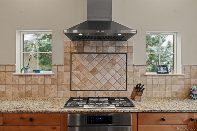 kitchen featuring wall chimney range hood, backsplash, light stone countertops, brown cabinets, and stainless steel appliances
