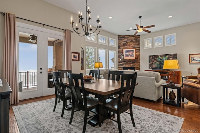 dining room featuring a stone fireplace, recessed lighting, ceiling fan with notable chandelier, dark wood-type flooring, and french doors