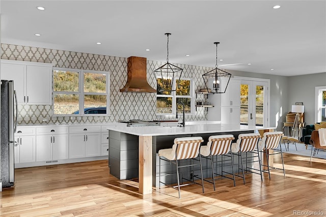 kitchen featuring wall chimney exhaust hood, stainless steel fridge, decorative light fixtures, a center island with sink, and white cabinets