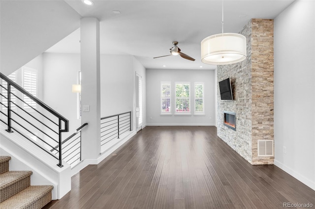 unfurnished living room featuring ceiling fan, dark wood-style floors, a stone fireplace, baseboards, and stairway