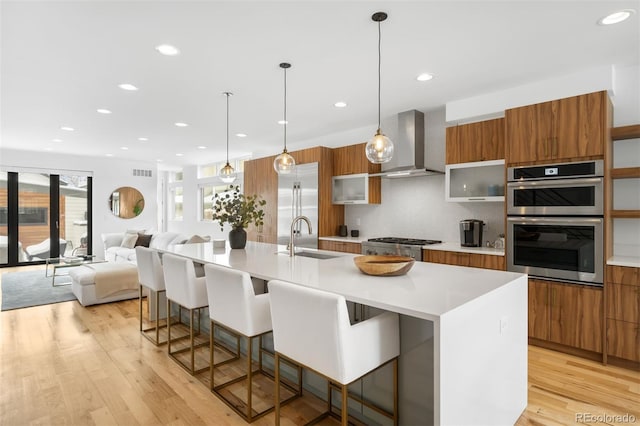 kitchen featuring appliances with stainless steel finishes, a kitchen island with sink, light countertops, wall chimney range hood, and a sink