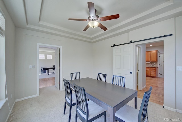 dining room with light carpet, a tray ceiling, a barn door, baseboards, and ceiling fan